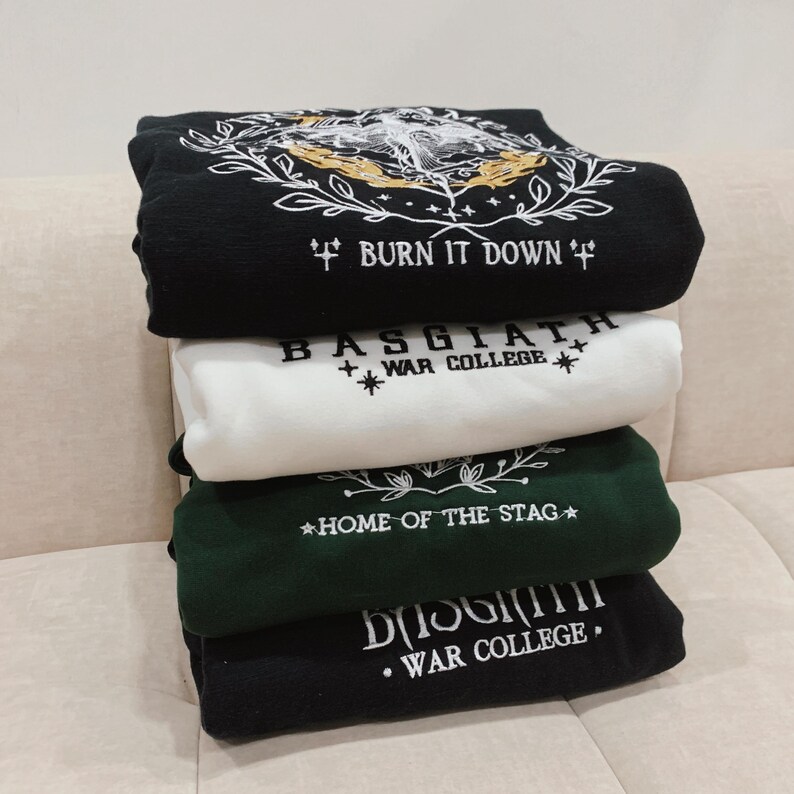 May include: A stack of four black and white sweatshirts with embroidered designs. The top sweatshirt has a black background with a white design of a hand holding a burning torch with the text "BURN IT DOWN". The second sweatshirt has a white background with the text "BASGIATH WAR COLLEGE" in black. The third sweatshirt has a green background with the text "HOME OF THE STAG" in white. The bottom sweatshirt has a black background with the text "BASGIATH WAR COLLEGE" in white.