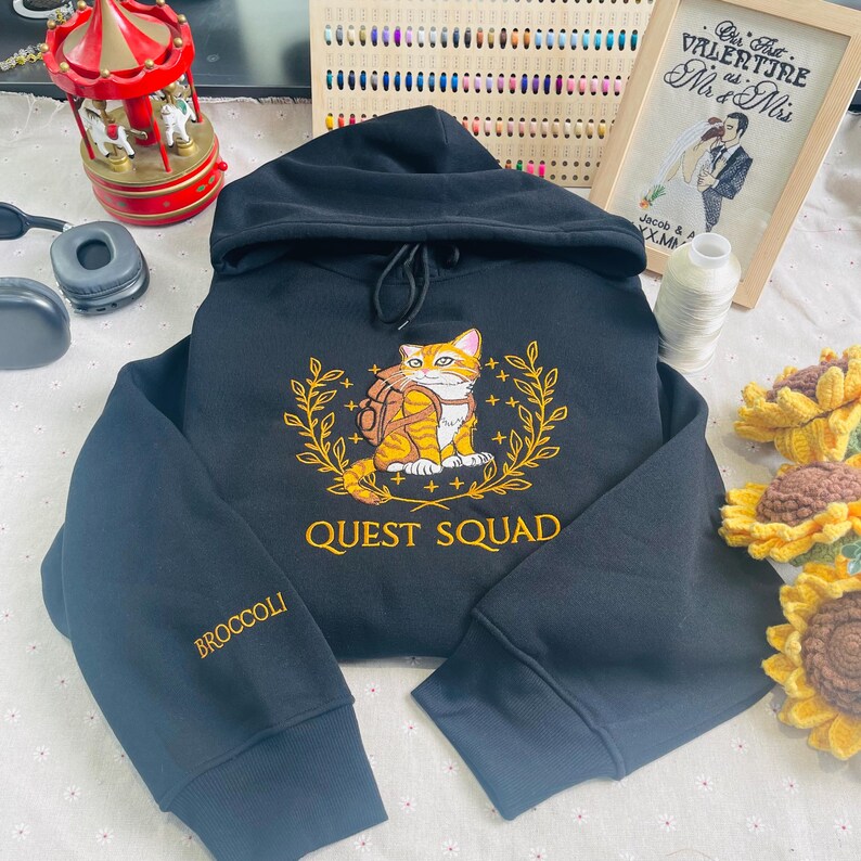 May include: Black hooded sweatshirt with a gold embroidered design of a cat wearing a backpack and a laurel wreath. The text "QUEST SQUAD" is embroidered below the design. The left sleeve has the word "BROCCOLI" embroidered in gold.