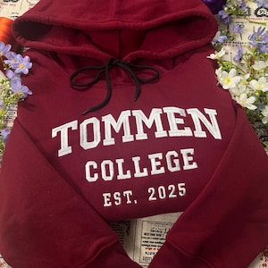 May include: A maroon hoodie with a drawstring hood. The front features the white text "TOMMEN COLLEGE EST. 2025". The hoodie is made of a soft material and is likely for casual wear. The design is simple and collegiate.