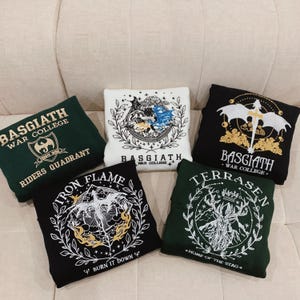 May include: Five different sweatshirts with embroidered designs. The sweatshirts are in various colors, including green, black, and white. The designs feature dragons, stags, and other mythical creatures. The text on the sweatshirts includes "Basgiath War College", "Iron Flame", "Burn It Down", "Terrasen", and "Home of the Stag".