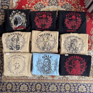 May include: A collection of eight beige, black and blue sweatshirts with different character designs from the video game Baldur's Gate. Each sweatshirt features a character's name and the words "Baldur's Gate".