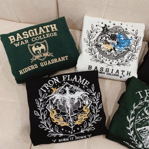 May include: Four green, black, and white sweatshirts with embroidered designs. The designs feature dragons, flames, and the text "BASGIATH WAR COLLEGE", "FOURTH WING", "RIDERS QUADRANT", "IRON FLAME", and "BURN IT DOWN".