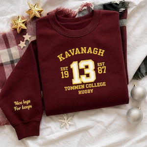 May include: A maroon sweatshirt with the text "Kavanagh 13 Est 1987 Tommen College Rugby" in yellow and white. The sleeve reads "Nice legs For keeps". The sweatshirt is surrounded by holiday decorations.