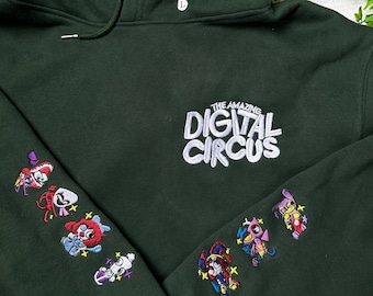 May include: A dark green hooded sweatshirt featuring the white embroidered text The Amazing Digital Circus on the chest. Both sleeves are decorated with colorful embroidered character designs and small star accents. A size L tag is visible.