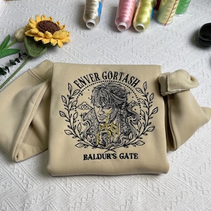 Könnte beinhalten: Ein beiger Pullover mit einem schwarzen Stickmuster, das ein Porträt einer Figur, den Text "Enver Gortash" und die Worte "Baldur's Gate" zeigt.