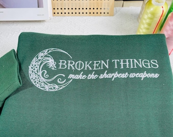 May include: A dark green sweatshirt with a white embroidered design. The design features a crescent moon with a stylized floral pattern and the text "BROKEN THINGS make the sharpest weapons".
