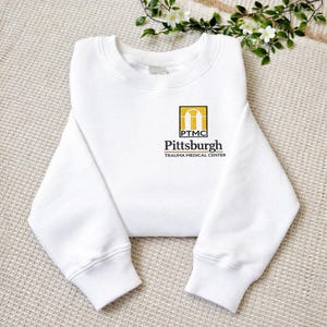 May include: A white crewneck sweatshirt featuring a small embroidered logo on the left chest. The logo includes a yellow arch icon above the text PTMC, followed by the words Pittsburgh and TRAUMA MEDICAL CENTER in black lettering.