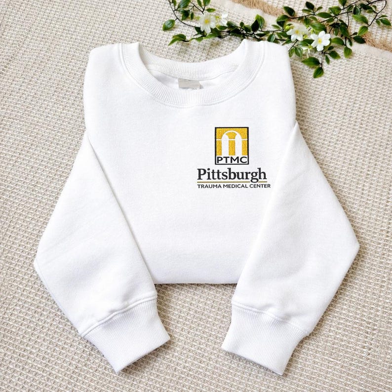 May include: A white crewneck sweatshirt featuring a small embroidered logo on the left chest. The logo includes a yellow arch icon above the text PTMC, followed by the words Pittsburgh and TRAUMA MEDICAL CENTER in black lettering.
