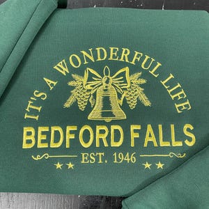 Pode incluir: Um moletom verde escuro com bordado dourado. O texto diz "IT'S A WONDERFUL LIFE" arqueado acima de "BEDFORD FALLS" e "EST. 1946". O design inclui um sino, um laço e estrelas.