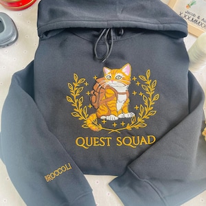 May include: Black hooded sweatshirt with a gold embroidered design of a cat wearing a backpack and a laurel wreath. The text "QUEST SQUAD" is embroidered below the cat. The left sleeve has the word "BROCCOLI" embroidered in gold.