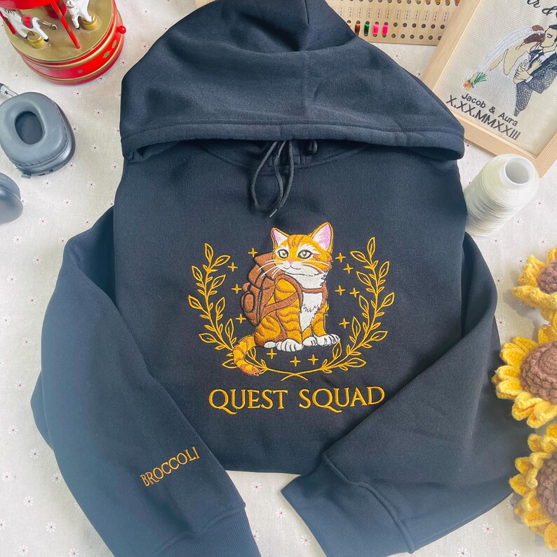 May include: Black hooded sweatshirt with an embroidered design of an orange tabby cat wearing a backpack and surrounded by a wreath of leaves. The text "QUEST SQUAD" is embroidered below the cat. The left sleeve has the word "BROCCOLI" embroidered on it.