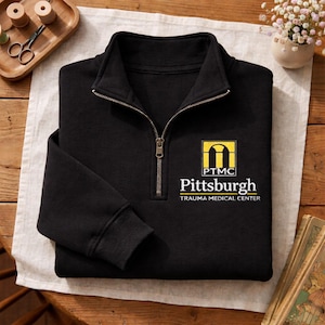 May include: A black quarter-zip pullover sweatshirt featuring a yellow and white embroidered logo on the left chest. The logo displays a stylized archway icon above the text PTMC, followed by the words Pittsburgh TRAUMA MEDICAL CENTER.