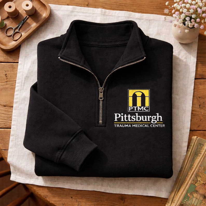 May include: A black quarter-zip pullover sweatshirt featuring a yellow and white embroidered logo on the left chest. The logo displays a stylized archway icon above the text PTMC, followed by the words Pittsburgh TRAUMA MEDICAL CENTER.