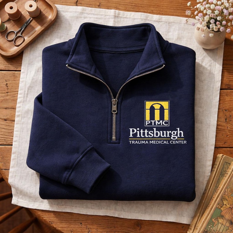 May include: A navy blue quarter-zip sweatshirt featuring a yellow and white logo on the left chest. The logo includes an arch graphic, the letters PTMC, and the text Pittsburgh TRAUMA MEDICAL CENTER printed in white and yellow lettering.
