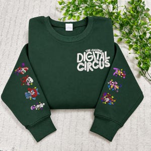 May include: A dark green crewneck sweatshirt featuring the white embroidered text The Amazing Digital Circus on the upper chest. Both long sleeves are decorated with small, colourful embroidered character graphics arranged in a vertical line.
