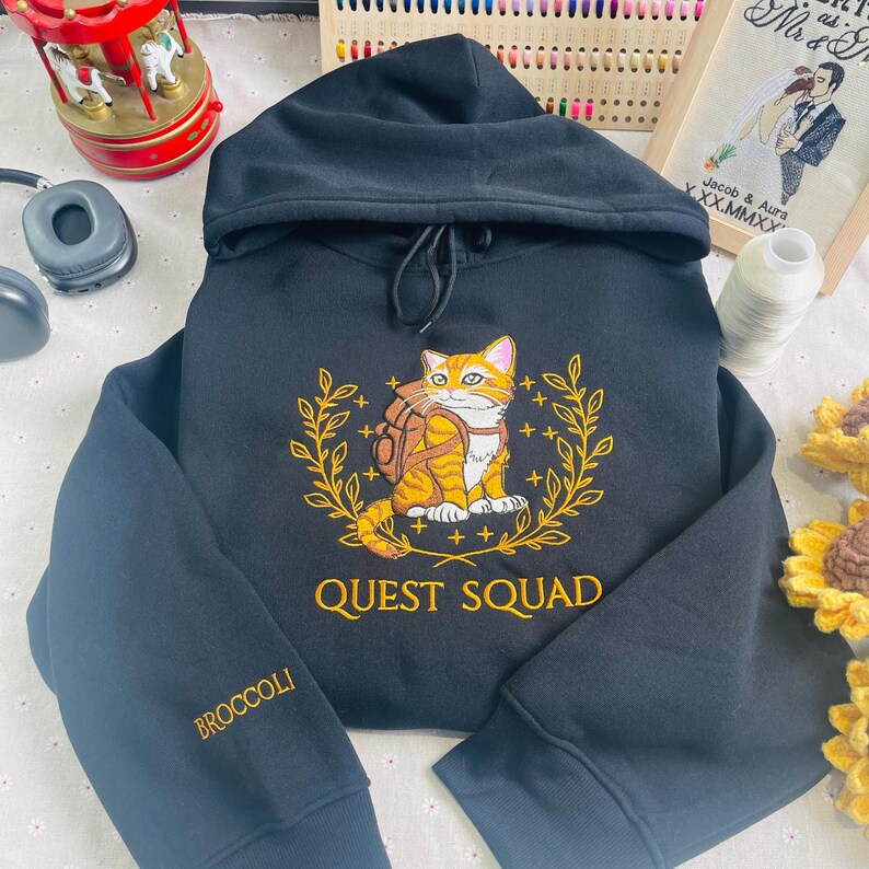 May include: Black hooded sweatshirt with a yellow embroidered design of a cat wearing a backpack and a laurel wreath. The text "QUEST SQUAD" is embroidered in yellow below the cat. The word "BROCCOLI" is embroidered in yellow on the left sleeve.