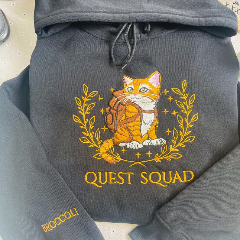 May include: Black hooded sweatshirt with an embroidered design of an orange tabby cat wearing a brown backpack and surrounded by gold leaves. The text "QUEST SQUAD" is embroidered below the cat. The word "BROCCOLI" is embroidered on the left sleeve.
