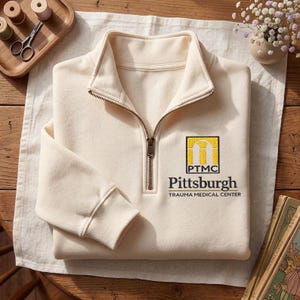 May include: A cream-colored quarter-zip pullover sweatshirt featuring an embroidered logo on the left chest. The logo includes a yellow arch graphic above the letters PTMC and the text Pittsburgh TRAUMA MEDICAL CENTER in black lettering.