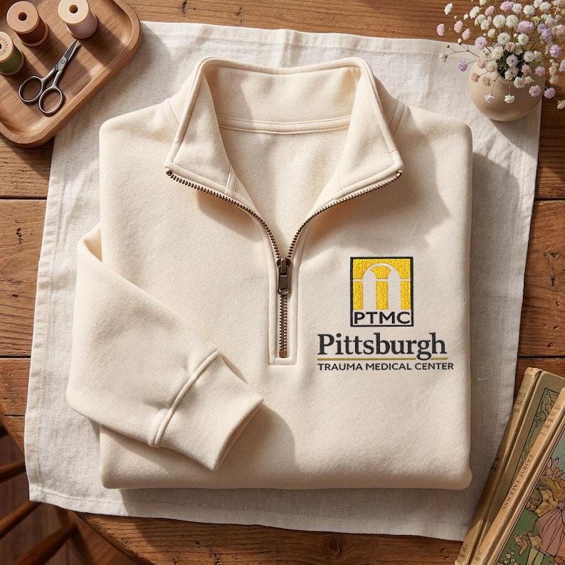 May include: A cream-colored quarter-zip pullover sweatshirt featuring an embroidered logo on the left chest. The logo includes a yellow arch graphic above the letters PTMC and the text Pittsburgh TRAUMA MEDICAL CENTER in black lettering.