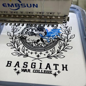 May include: A white fabric with black embroidery of a wreath, a celestial design, and the text "BASGIATH WAR COLLEGE". A blue dragon is embroidered on top of the celestial design. The fabric is being embroidered by a white embroidery machine.