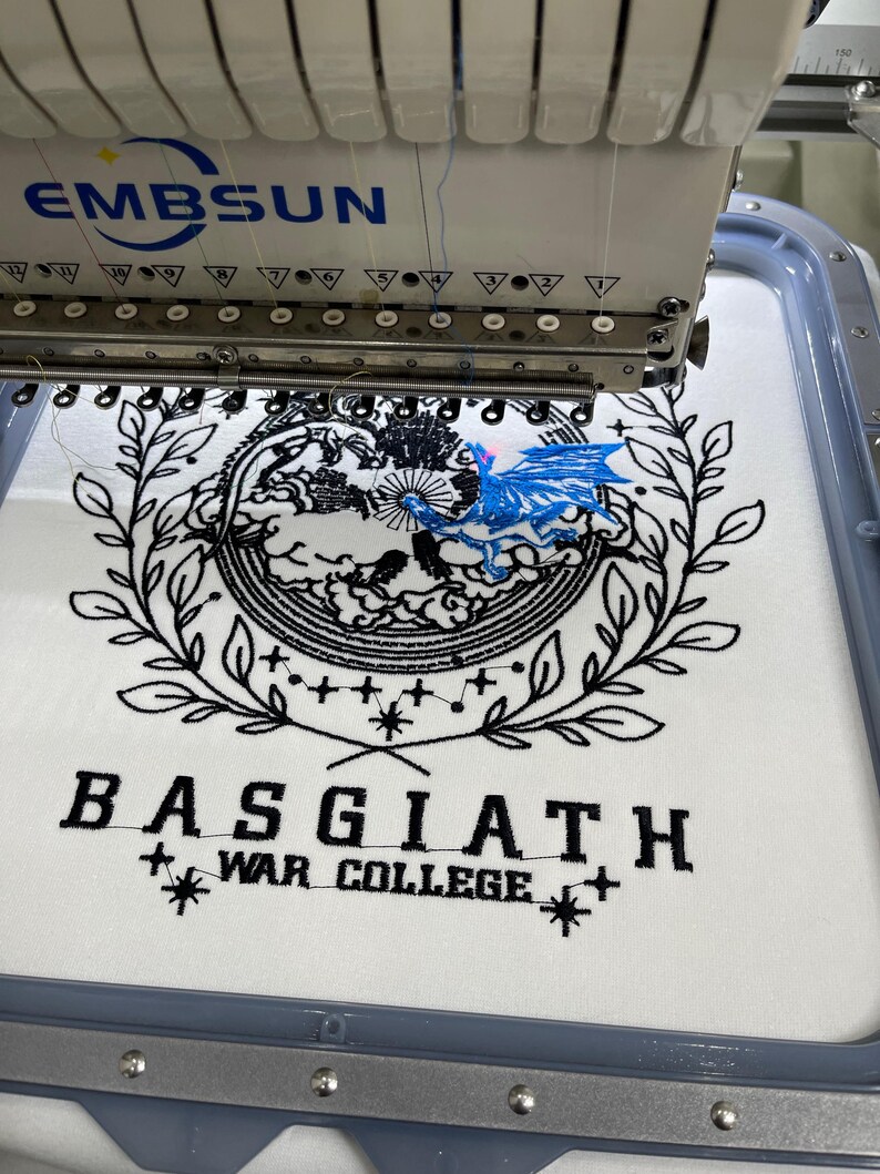 May include: A white fabric with black embroidery of a wreath, a celestial design, and the text "BASGIATH WAR COLLEGE". A blue dragon is embroidered on top of the celestial design. The fabric is being embroidered by a white embroidery machine.