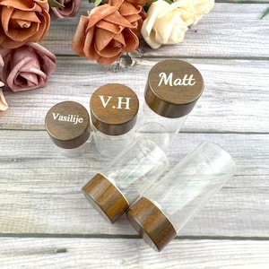 Puede incluir: Frascos de vidrio transparente con tapas de madera. Las tapas están grabadas con nombres e iniciales: "Matt", "Vasilije" y "V.H". Los frascos están dispuestos sobre una superficie clara con vetas de madera, con flores en el fondo.