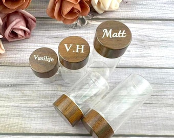 May include: Clear glass jars with wooden lids. The lids are engraved with names and initials: "Matt", "Vasilije", and "V.H". The jars are arranged on a light wood-grain surface, with flowers in the background.