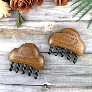 Puede incluir: Dos peines de pelo de madera con forma de nube y dientes negros. Un peine tiene dos corazones grabados, el otro tiene el texto "Johnny Best Man" en un marco decorativo. Los peines están sobre una superficie de madera clara.