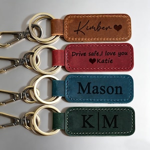 May include: Four personalized leather keychains in brown, red, teal, and green. Each keychain features a metal ring and clasp. The brown keychain is engraved with the name "Kimber" and a heart. The red keychain says "Drive safe, I love you Katie" with a heart. The teal keychain says "Mason". The green keychain has the initials "K|M".