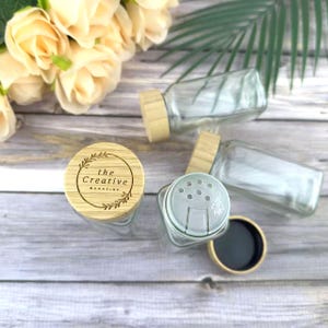 Peut inclure: Des pots à épices en verre transparent avec des couvercles en bambou. Un pot a un couvercle avec le texte "the Creative Magazine". Les pots sont rectangulaires avec un dessus de shaker et un couvercle en bambou.