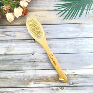 Puede incluir: Un cepillo seco de madera clara con mango largo y cerdas naturales. El mango está grabado con el nombre "Simon". El cepillo tiene un lazo blanco para colgar. Diseñado para el cuidado corporal.