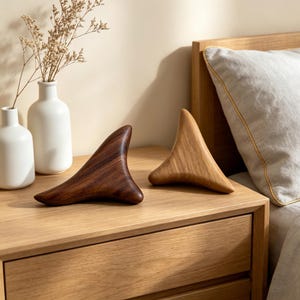 May include: Two wooden massage tools, one dark brown and one light brown, sit on a wooden nightstand. The tools have a curved, triangular shape. Two white vases with dried flowers are in the background.