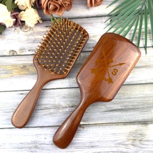 Könnte beinhalten: Zwei Holzbürsten mit dunkelbraunem Finish. Eine Bürste hat Holzborsten, die andere ein eingraviertes Design aus Pfeilen und dem Buchstaben "S". Beide Bürsten haben einen rechteckigen Kopf und einen gebogenen Griff.