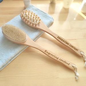 Puede incluir: Dos cepillos de baño de madera con cerdas naturales y un mango de cuerda anudada. Los mangos están grabados con el texto "Engraving content".