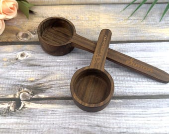 May include: Two wooden coffee scoops with a dark brown finish are displayed on a light wood surface. The word "Simon" is engraved on the handle of each scoop. The scoops are designed with a round bowl and a long handle.