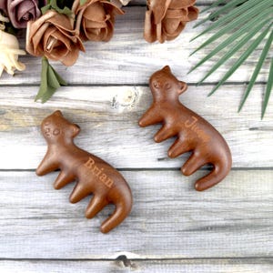 Könnte beinhalten: Zwei Massage-Tools in Bärenform aus Holz mit den eingravierten Namen "Brian" und "Juan". Die Tools sind warmbraun und liegen auf einer hellgrauen Holzoberfläche, mit floralen und Blatt-Akzenten im Hintergrund.