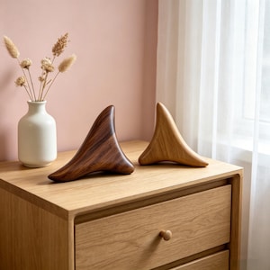 May include: Two wooden sculptures with a curved, abstract design sit on a light wood nightstand. One is a dark brown, the other a light tan. A small white vase with dried flowers is to the left. The background is a soft pink wall and a sheer white curtain.