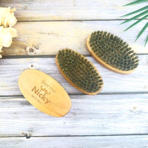 Könnte beinhalten: Drei Bartbürsten aus Holz mit dunklen Borsten. Eine Bürste ist mit "Best man Nicky 09.12.2021" und einem Schnurrbart-Symbol graviert. Die Bürsten sind oval und aus hellem Holz.