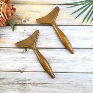May include: Two wooden massage tools with a curved head and a long handle. The handles have the name "Sharon" engraved in an orange color. The tools are made of a light brown wood and are displayed on a light wood surface.