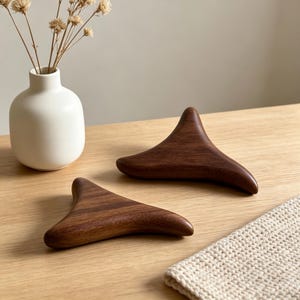 May include: Two dark brown wooden gua sha tools are displayed on a light wood surface. The tools have a smooth, curved shape. A small white vase with dried flowers is in the background, along with a cream-colored knitted cloth.