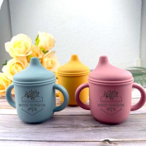 May include: Three colorful silicone baby cups with handles. One is light blue, one is yellow, and one is pink. Each cup has a lid and the text "HANDCRAFTED BY WOOD SURGEON" printed on the front. The cups are designed for infants and toddlers.