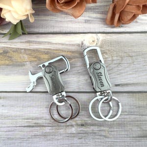 May include: Two silver keychains with personalized nameplates. One keychain has "Simon" engraved, and the other has "Steven". Both have a bottle opener and key rings. The keychains are on a wooden surface.