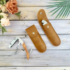 May include: A corkscrew with a wooden handle engraved with the name "James", and two brown leather cases, one open and one closed, each holding a silver bottle opener. The items are on a light wood-grain surface.