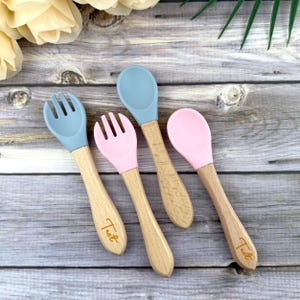 May include: Four baby utensils: two forks and two spoons. The fork and spoon heads are light blue and pink, attached to wooden handles. The word "Test" is printed in gold on the handles. The utensils are arranged on a wooden surface.