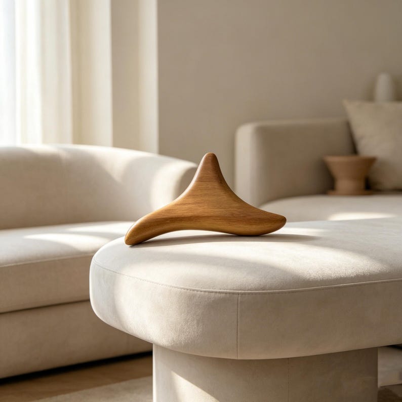 May include: A wooden massage tool with a curved, wing-like shape rests on a cream-colored ottoman. The tool is made of light brown wood and has a smooth, polished finish. The background features a neutral-toned living room with a sofa and natural light.
