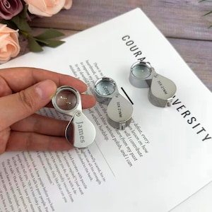 May include: Silver folding magnifying glasses with a 30X-21MM lens are shown on white paper with text. One magnifier is engraved with the name "James". The background includes flowers.