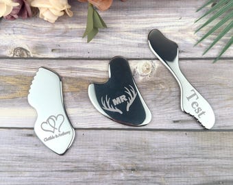 May include: Three silver-colored massage tools on a wooden surface. One tool is a spatula shape with the word "Test" engraved. Another is a heart-shaped tool with "Clothilde & Anthony" engraved. The third is a dark gray tool with "MR" and antlers engraved.