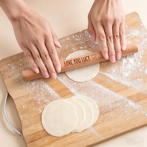 May include: A wooden rolling pin with the text "LOVE YOU LUCY" is used to flatten dough on a wooden cutting board. Several round pieces of dough are visible. Flour dusts the board.
