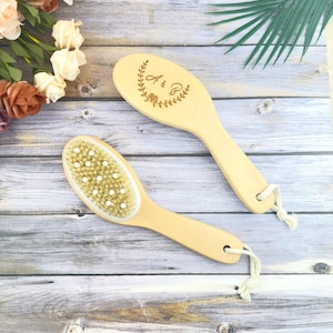 May include: Two wooden body brushes with light-colored bristles and handles. One brush has a decorative floral design with the letters "A & B". Both brushes have a loop for hanging. The brushes are on a light wood-grain surface.