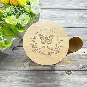 May include: A round wooden coaster with a butterfly design and the initials "A.C." engraved in the center. The coaster is surrounded by a floral design.